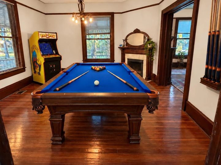 The game room: a pool table with blue felt under a brass chandelier, and a Ms. Pac-Man arcade cabinet by the window.